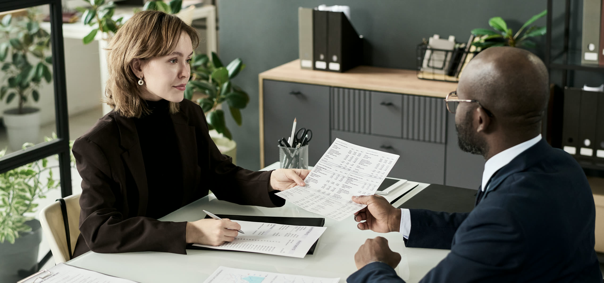 A solicitor at a desk in a town-centre office, a paper matter file open beside a laptop, deed boxes and lever-arch files on the shelving behind.