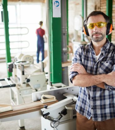 Waist up portrait of confident mature carpenter standing with arms crossed in workshop, copy space